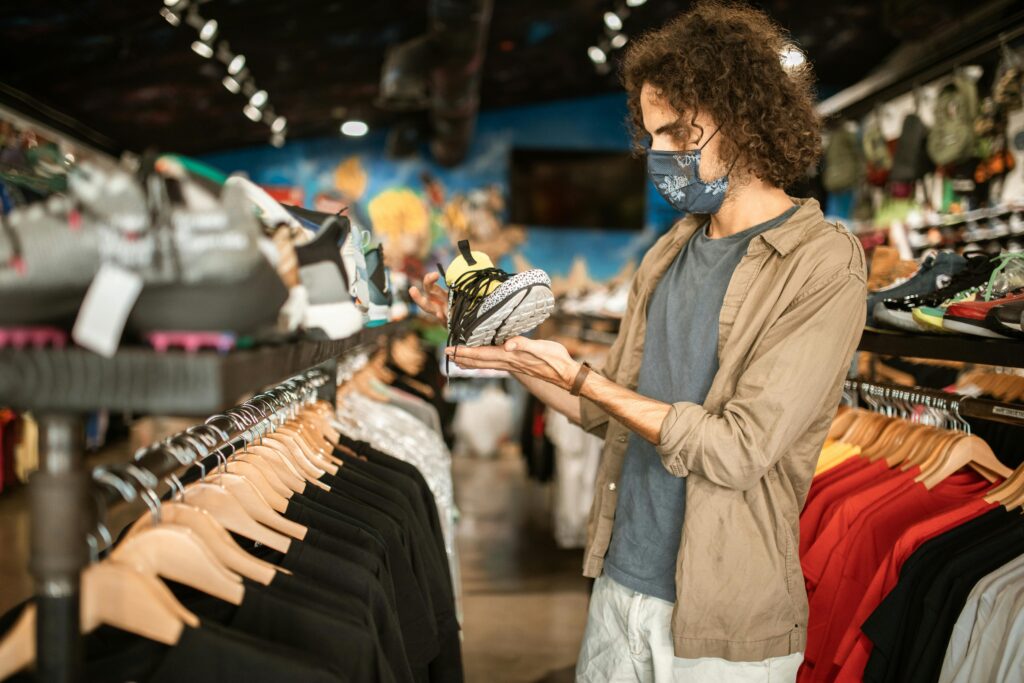 A man wearing a face mask checks sneakers in a stylish clothing store, surrounded by trendy apparel.