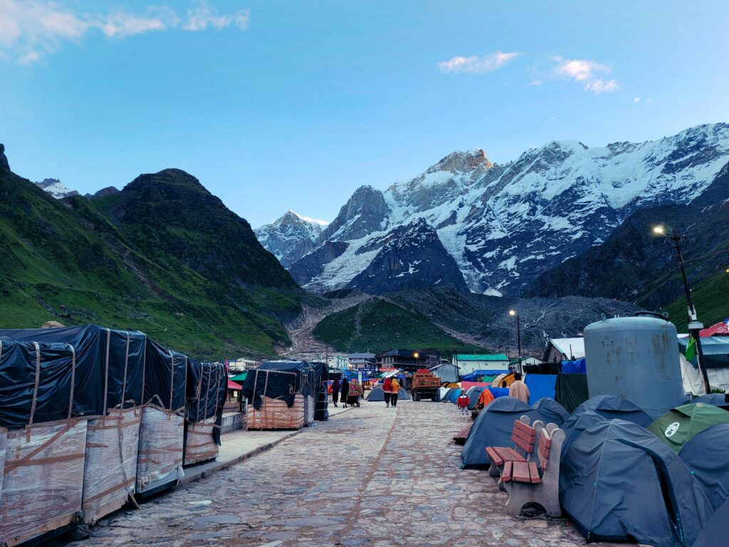 Picturesque Kedarnath setting with majestic snow-covered mountains and tents along a pathway.